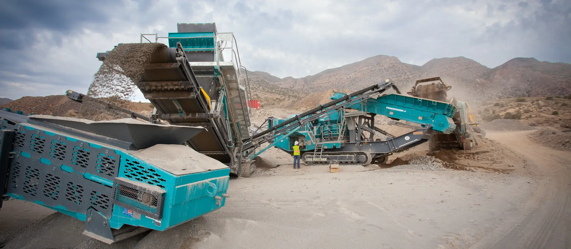 A tracked crushing and screening plant working an ore stockpile below desert mountains
